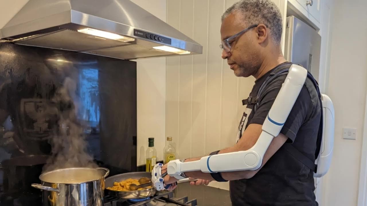 Portant l'exosquelette, un homme cuisine à une cuisinière à gaz — les biellettes accompagnent sa main atteinte qui manie un ustensile dans la poêle, l'autre main tenant le manche, tandis que la vapeur monte de la casserole voisine